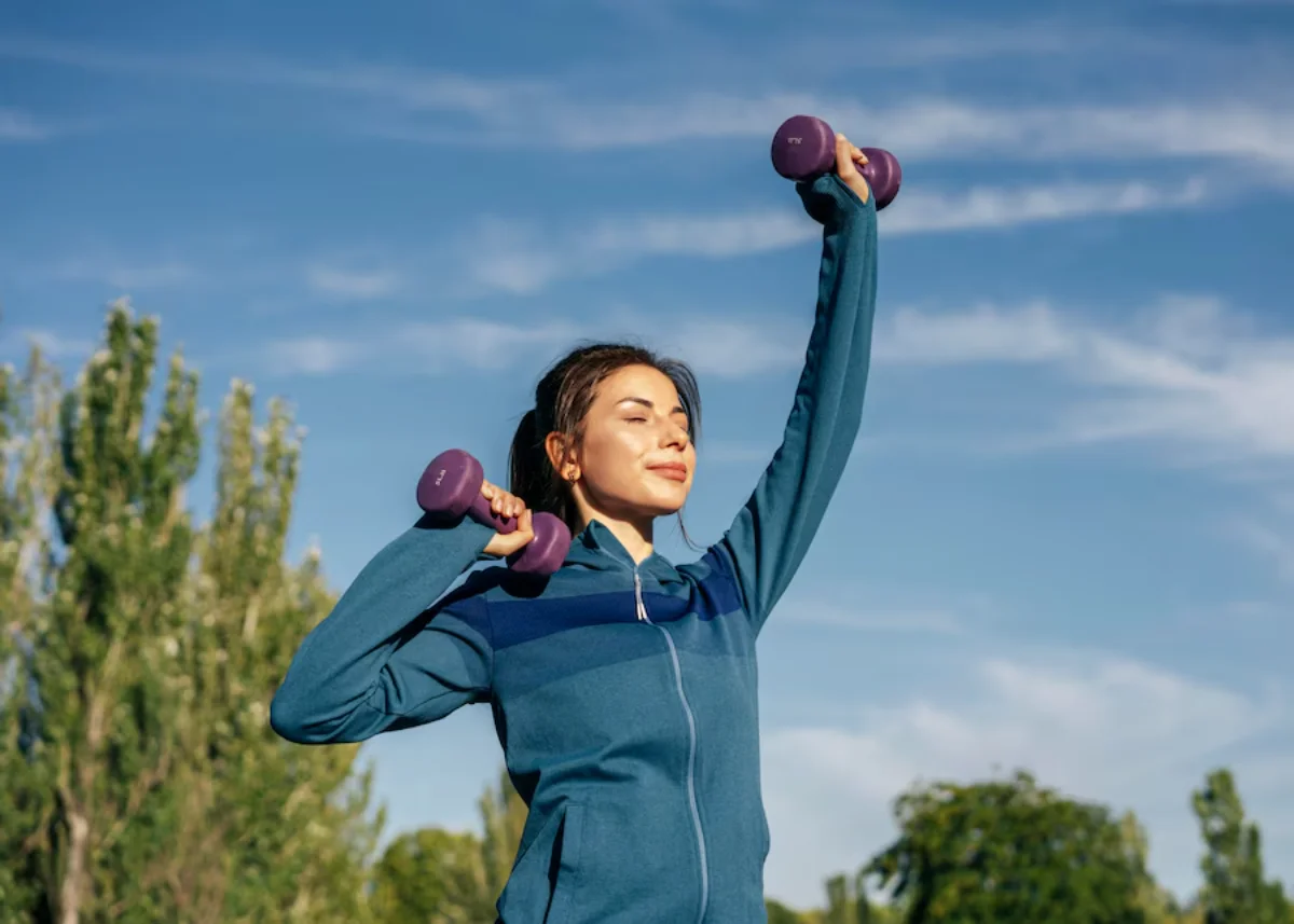A woman exercises outdoors, lifting weights under a clear blue sky.