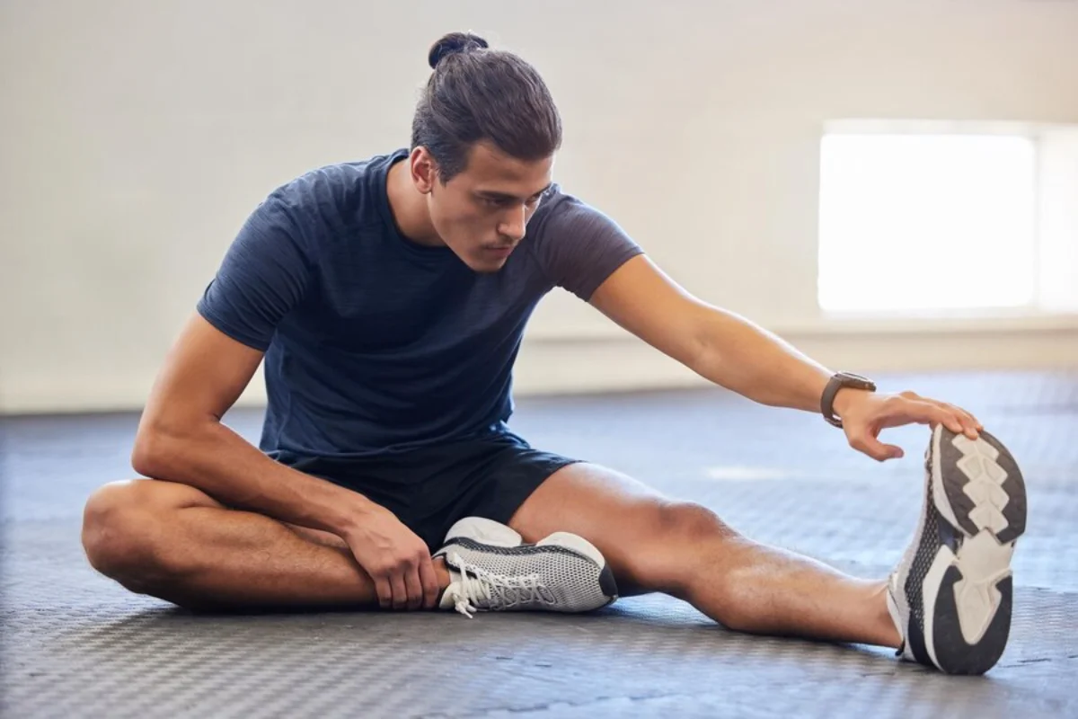Young man stretching his leg in a gym, touching his toes while sitting on the floor.