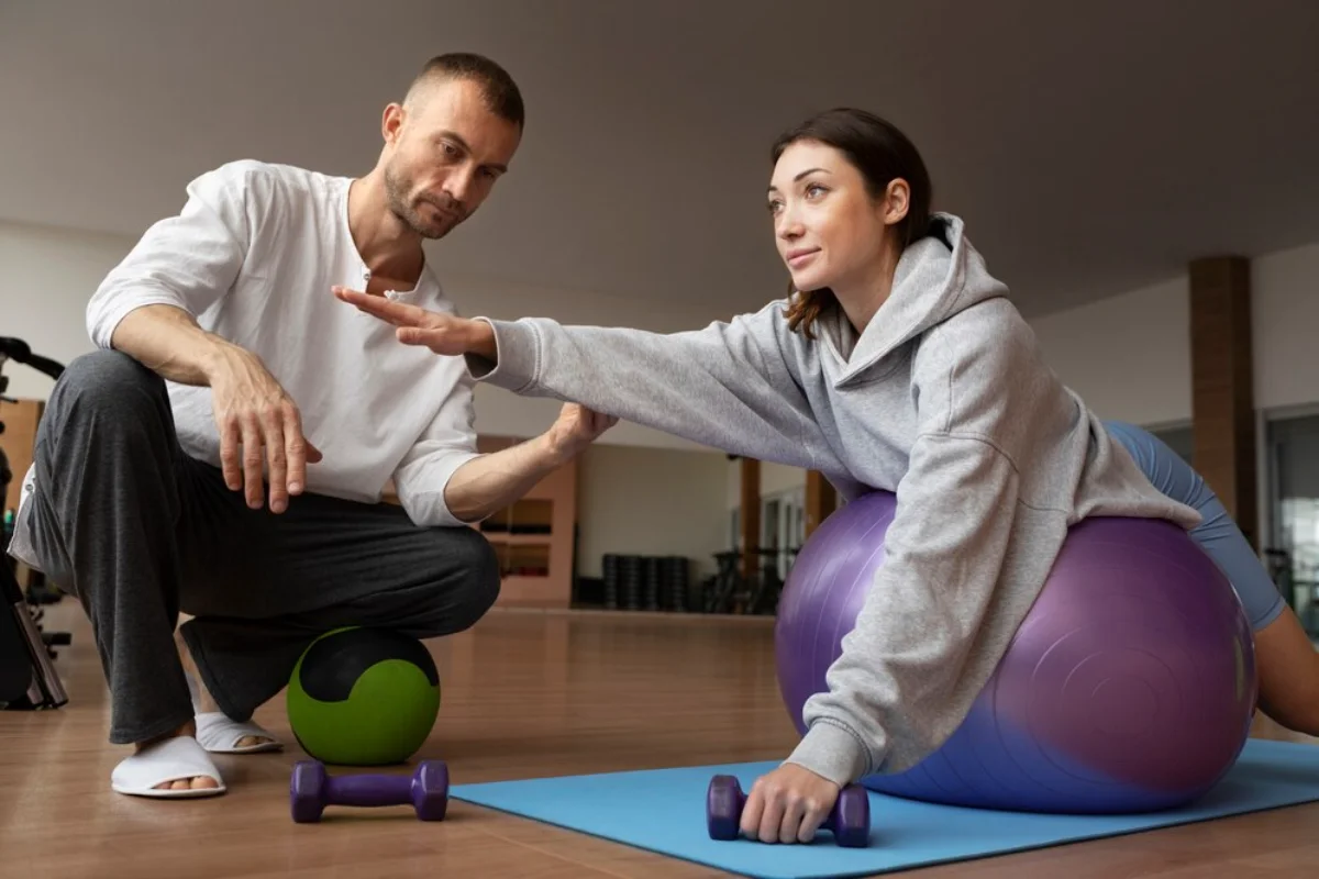 A man instructing a woman exercising with a stability ball in a gym.