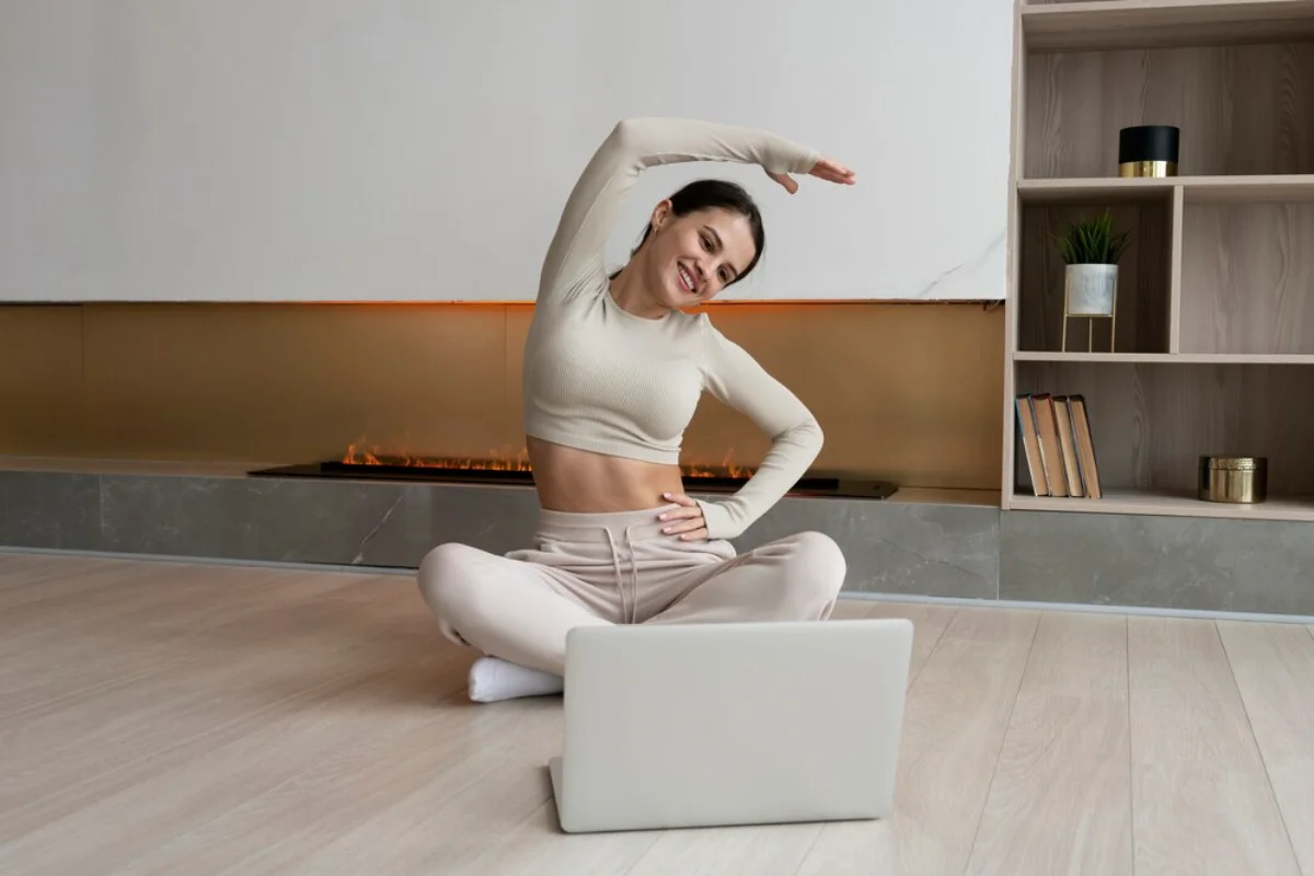 Woman stretching beside a laptop on the floor, in a cozy, modern room with a fireplace.