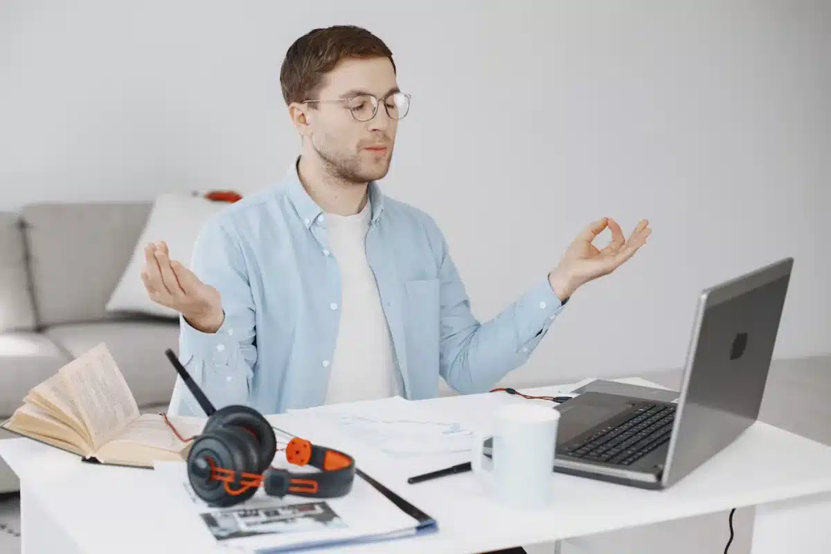 Man meditating at desk with laptop, books, and headphones.