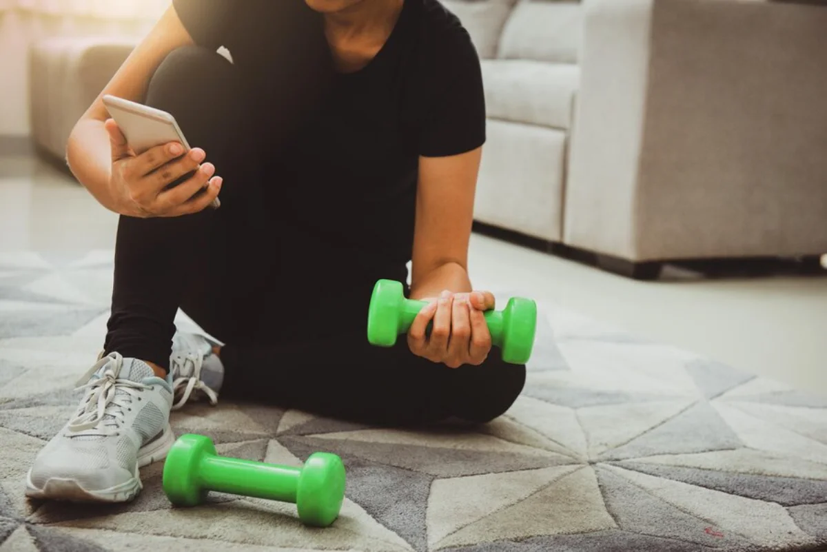 Person sitting on floor holding a dumbbell and smartphone, with another dumbbell nearby, in a home setting.