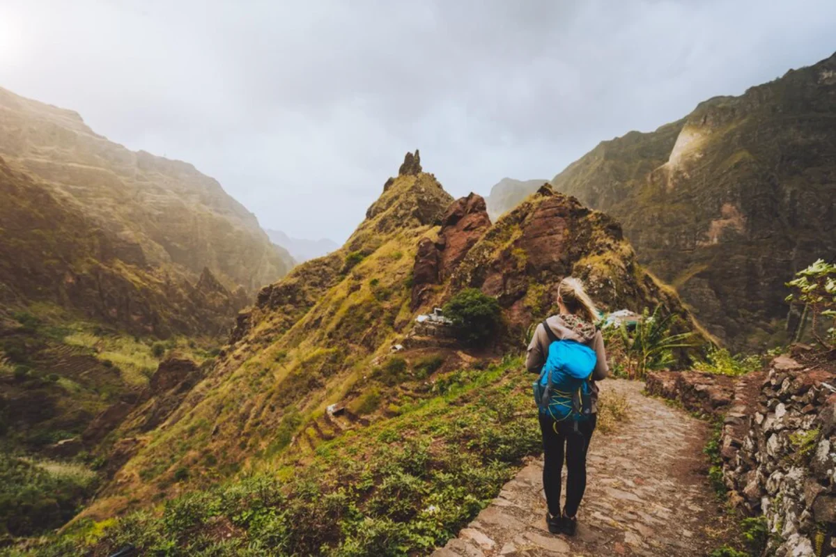 A hiker with a blue backpack walks a mountain path, scenic rocky peaks and misty hills surrounding.