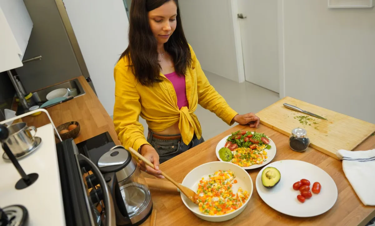 A woman in a yellow jacket serves various healthy dishes in a modern kitchen.