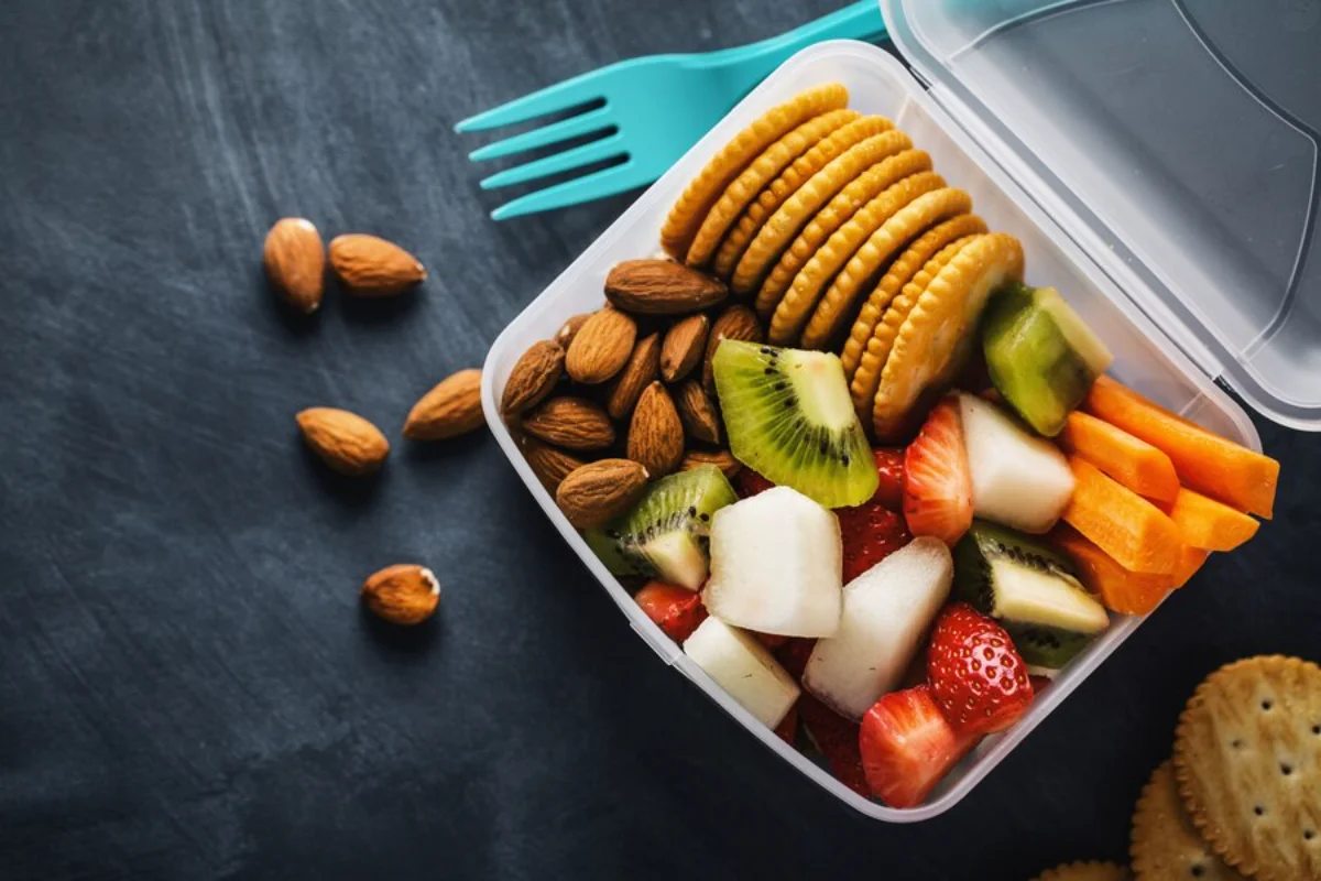 A lunch box filled with fruits, vegetables, nuts, and crackers on a dark surface.