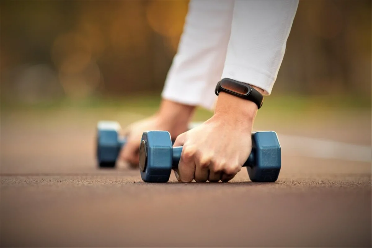 Person wearing a fitness tracker preparing to lift blue dumbbells on a path outdoors.