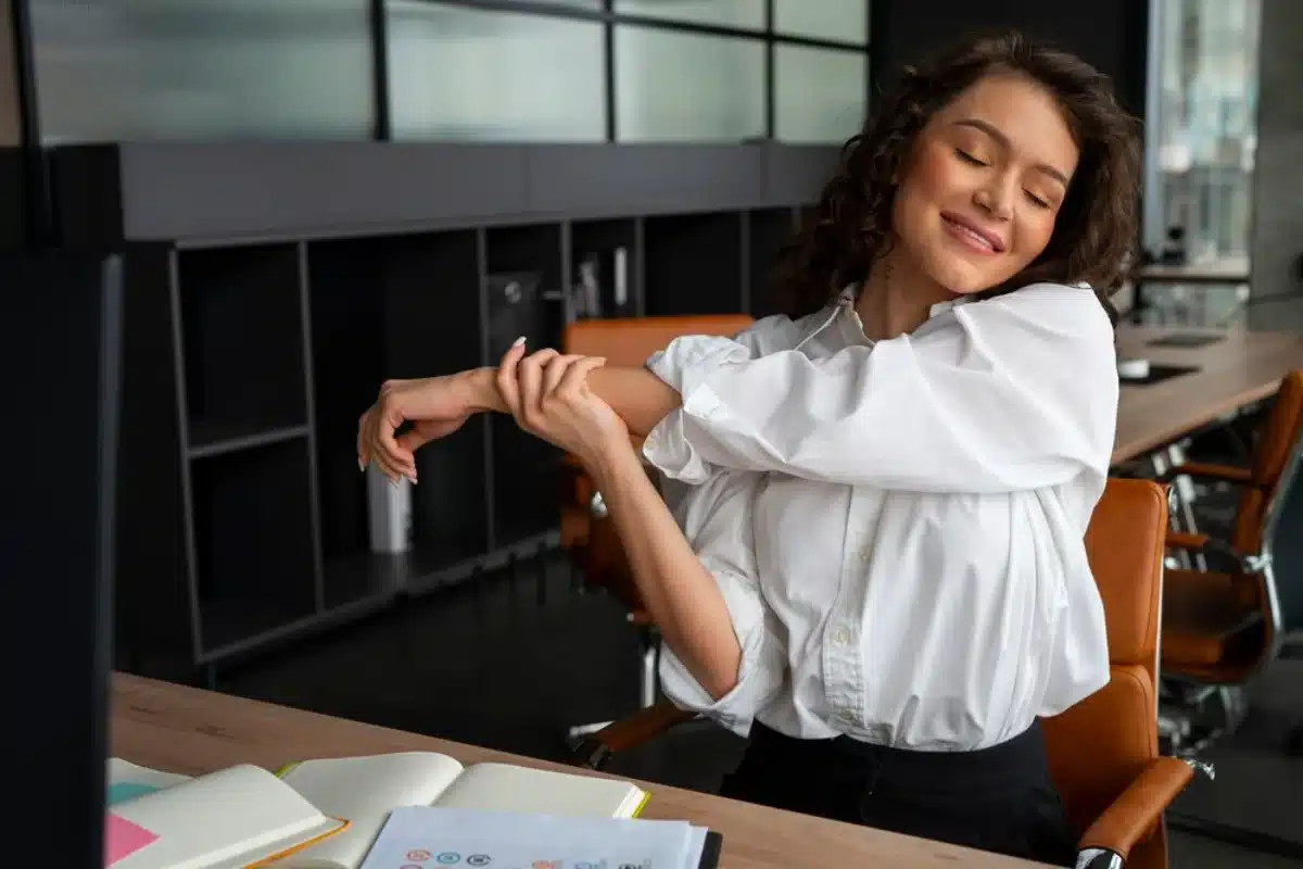 Woman stretching arms at desk in office, smiling with eyes closed, wearing a white shirt.