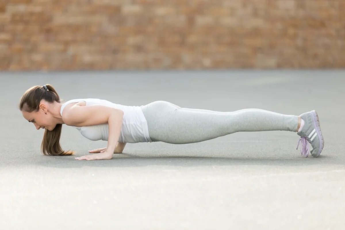 Woman doing a push-up outdoors on concrete, wearing white workout gear and grey trainers.
