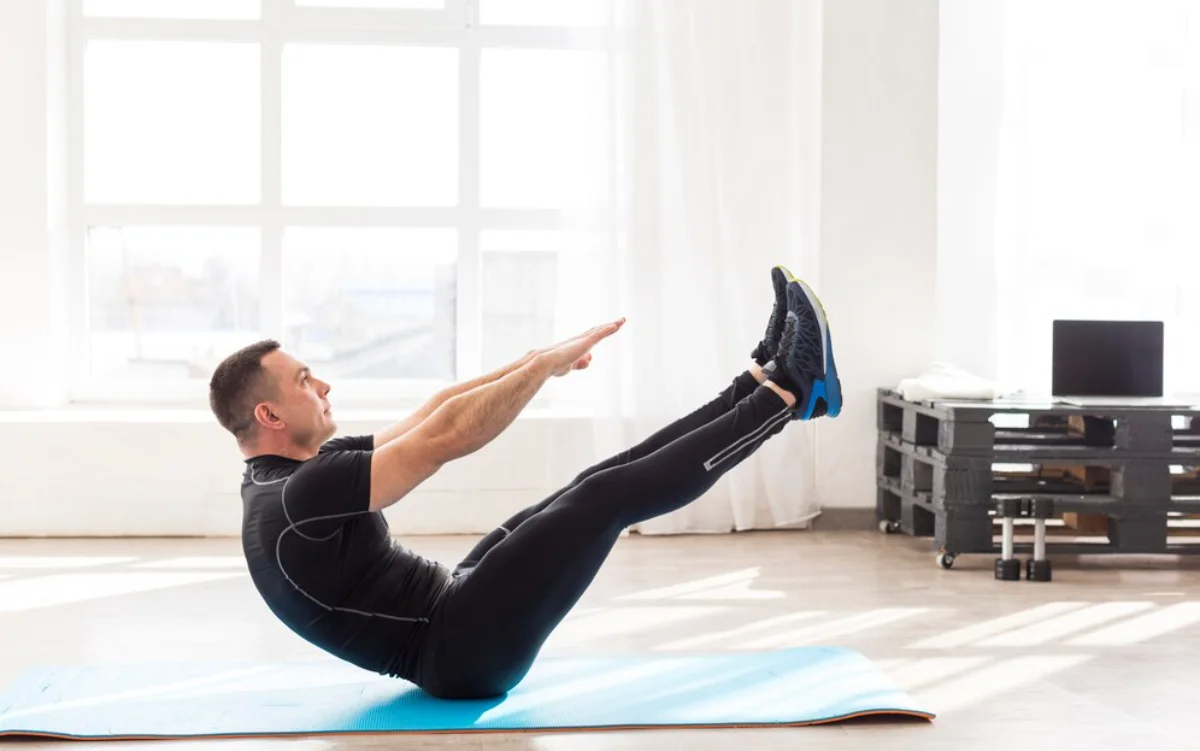 Man performing a V-sit abdominal exercise on a blue mat in a bright room with large windows.
