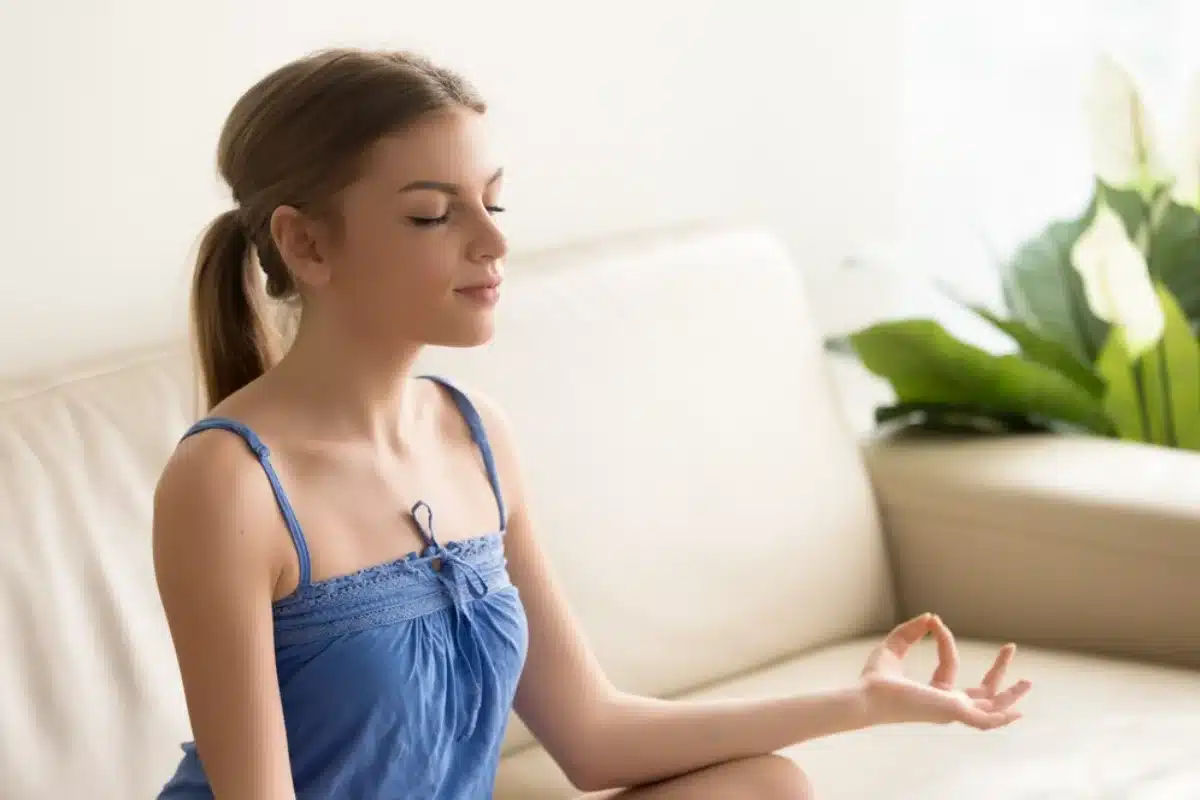 Young woman meditating peacefully indoors, sitting on floor with eyes closed and hands in mudra gesture.