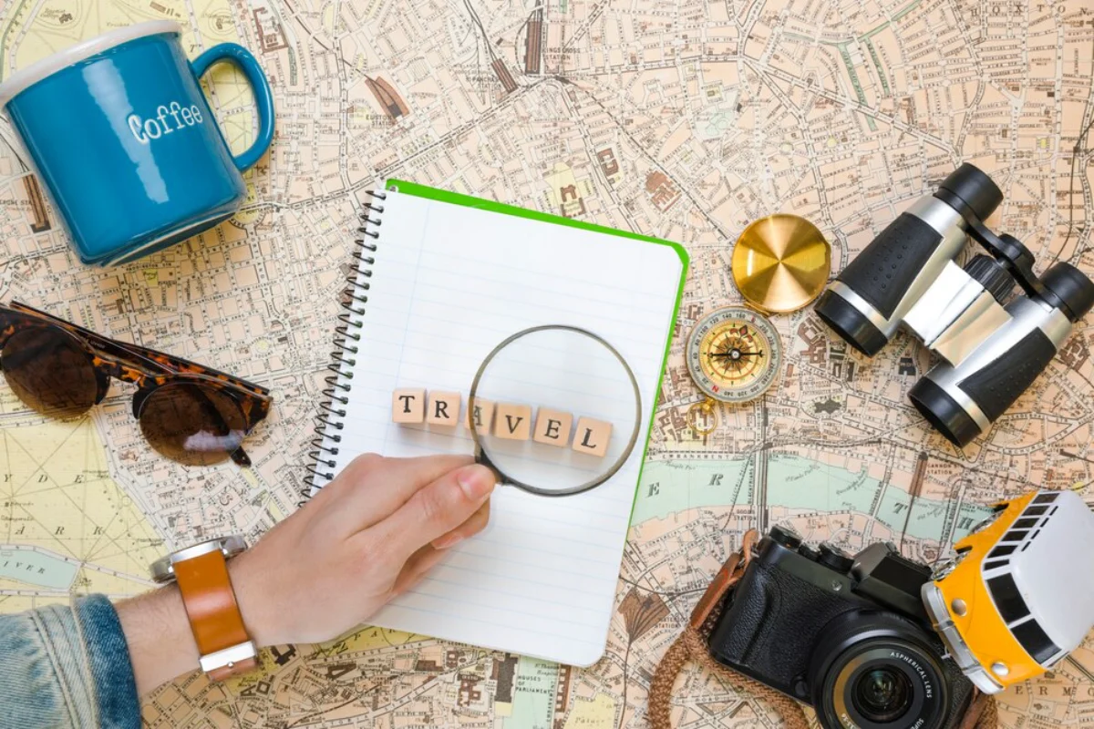 Travel planning setup with map, notebook, magnifying glass, binoculars, and camera on a table.