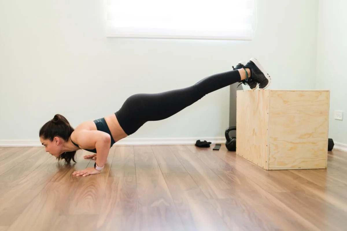Woman performing an elevated plank exercise with feet on a wooden box in a home gym setting.