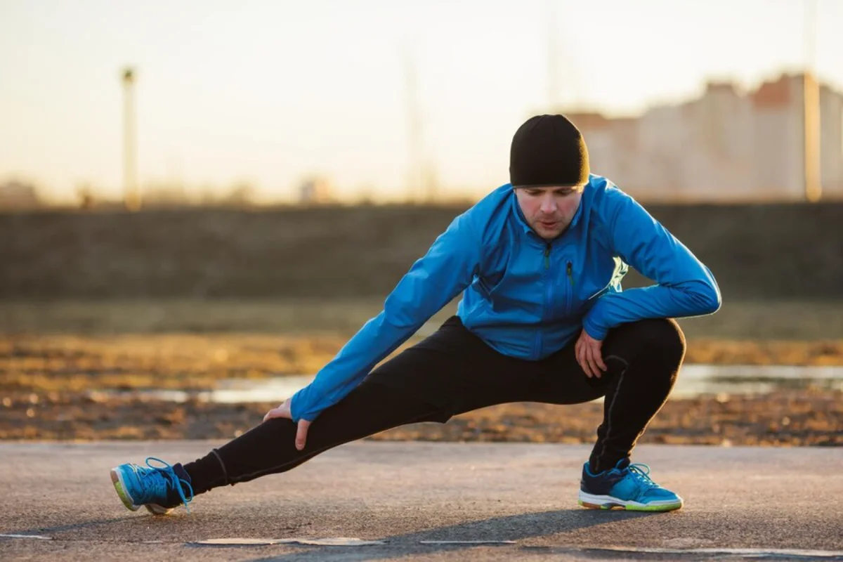 Man in blue jacket and black hat stretching outdoors at sunset.