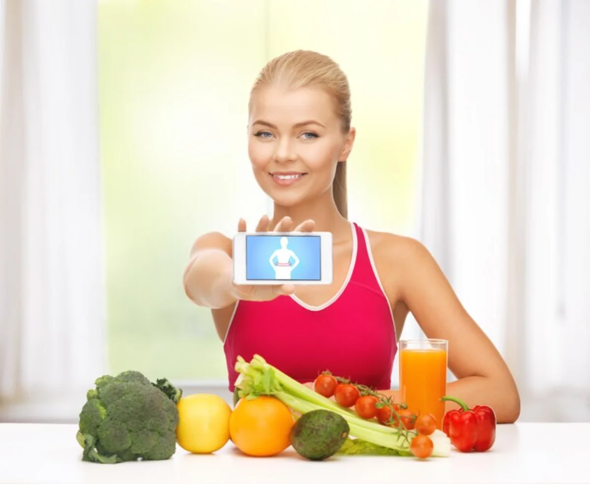 Young woman showing a health app on her phone, surrounded by fresh vegetables and a glass of juice.
