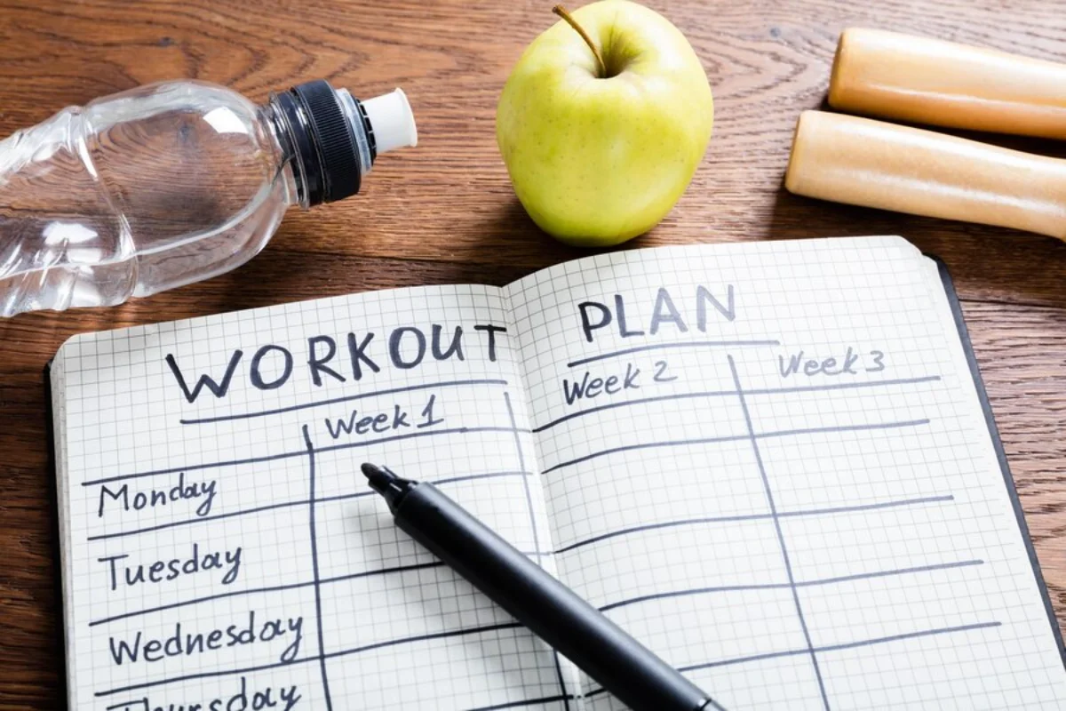 Notebook with handwritten workout plan, fresh apple, water bottle, and dumbbells on wooden table.
