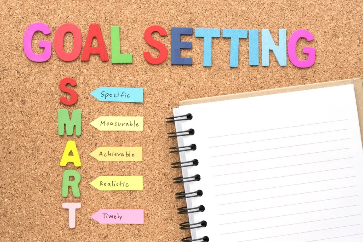 Colourful GOAL SETTING and SMART acronyms on corkboard with notepad, depicting goal planning concepts.
