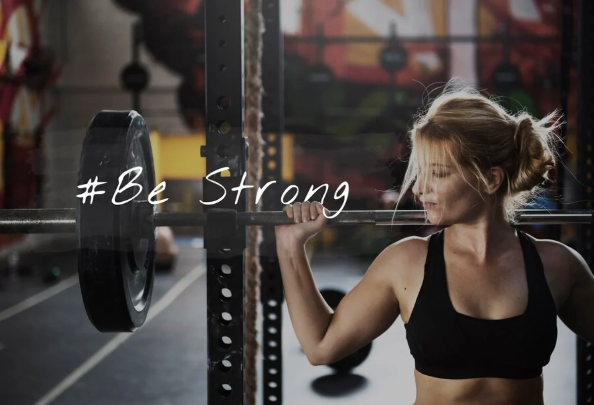 Woman lifting weights at a gym with #Be Strong text overlay.