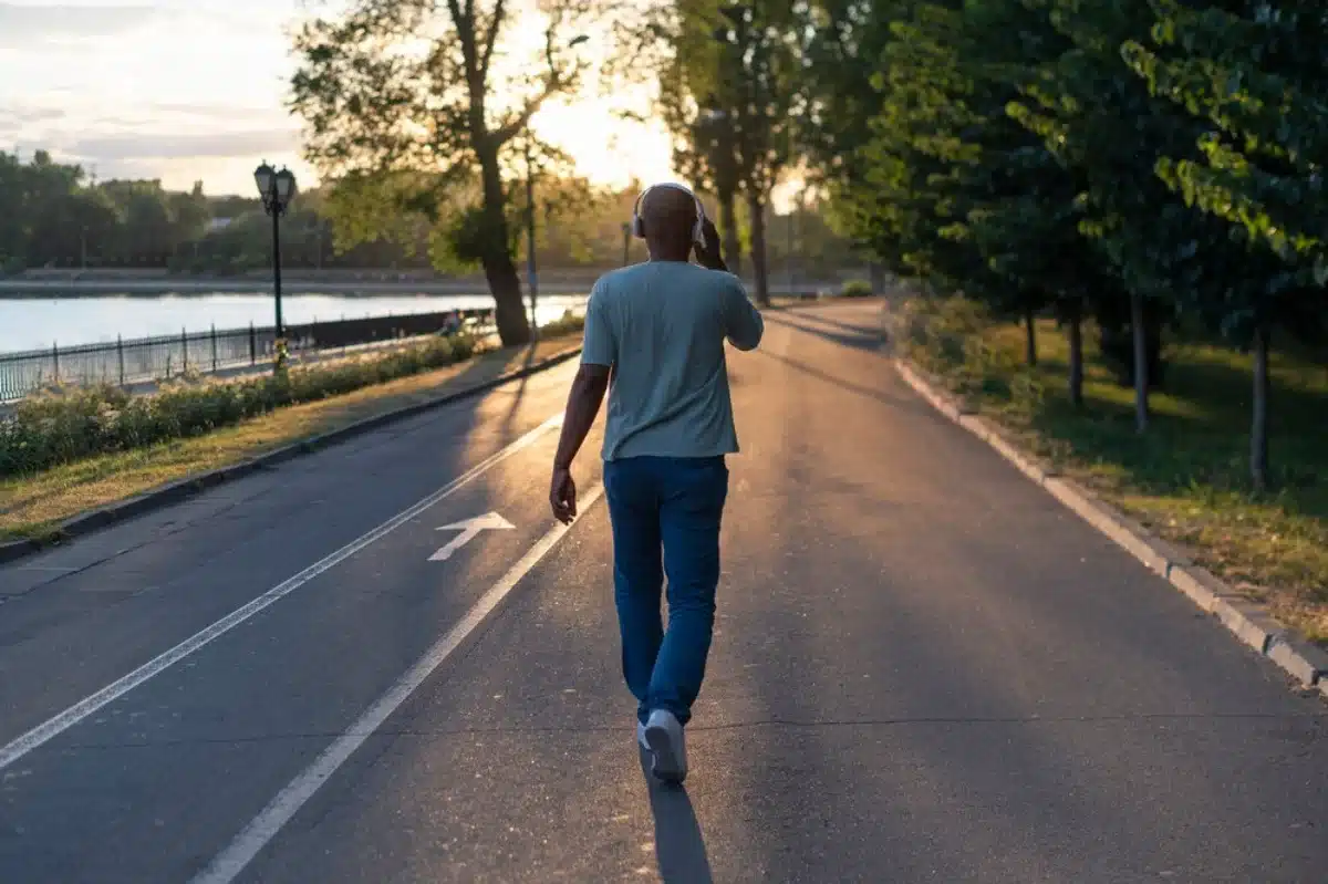 Man walking along a tree-lined park path near a river at sunset, wearing headphones.