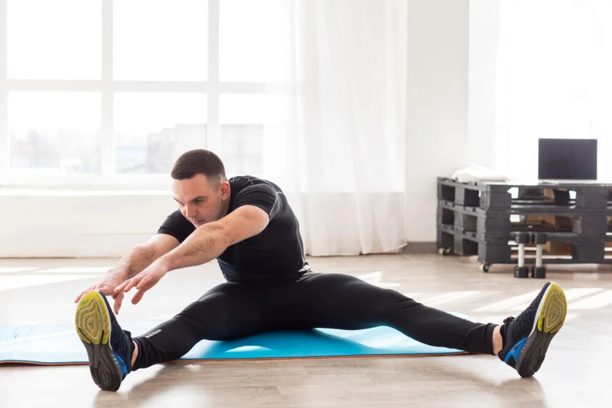 Man stretching on a mat in a bright room, reaching towards his outstretched foot.