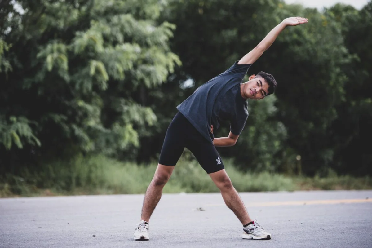 Young man doing a side stretch on a road, surrounded by lush greenery.