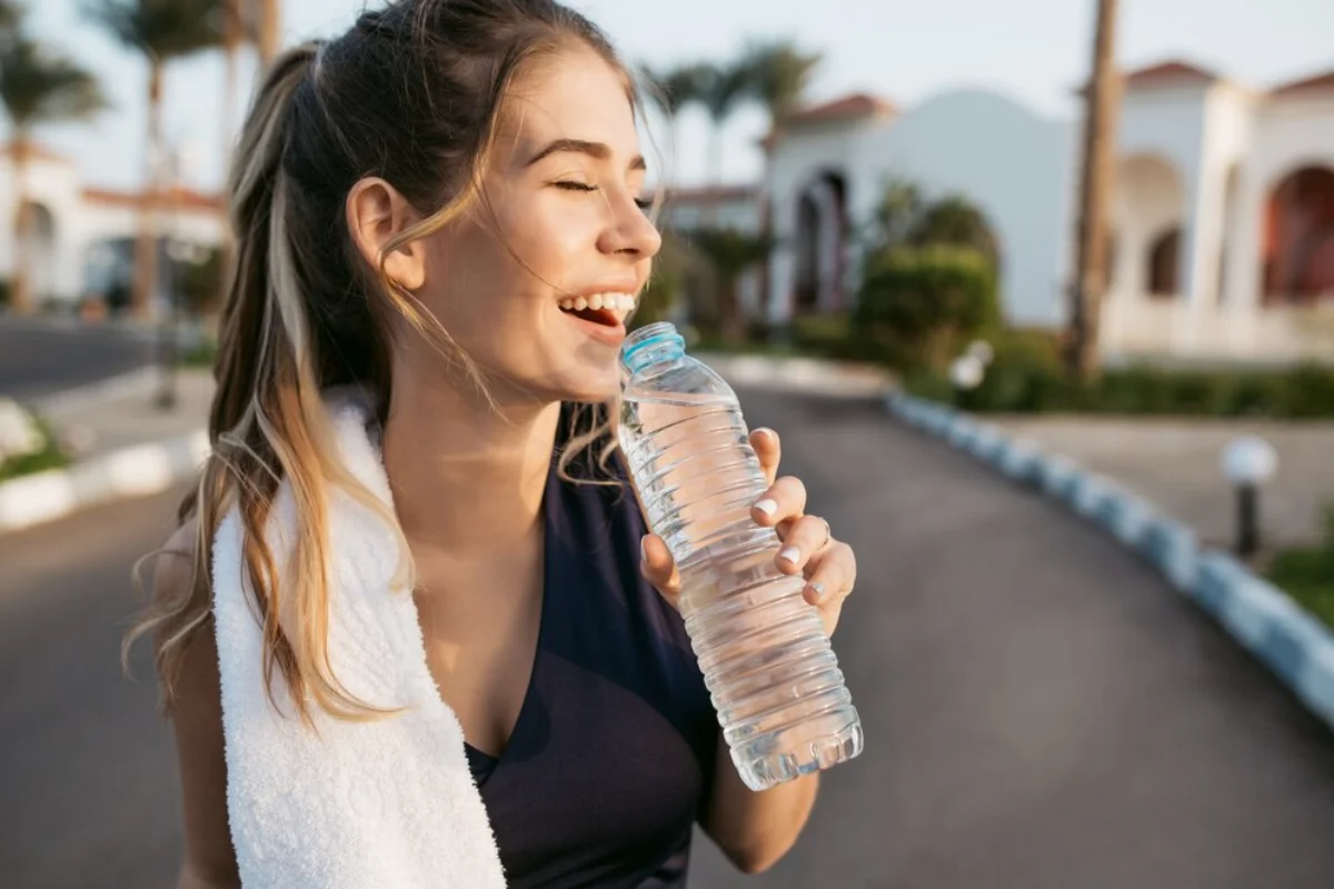 Young woman smiling and drinking from a water bottle outdoors.