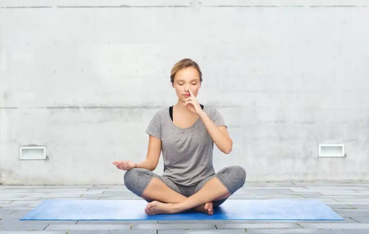 Woman practicing yoga in a cross-legged pose on a blue mat outdoors, against a grey wall.