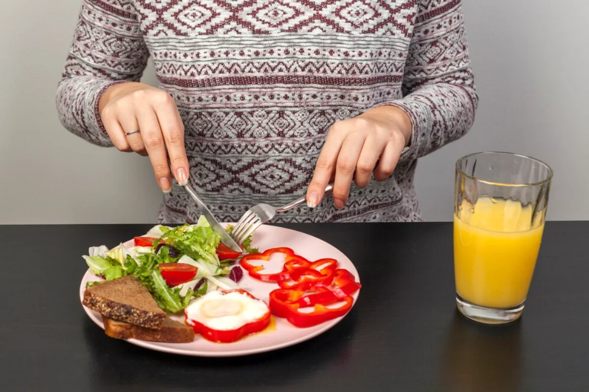 Person eating a healthy meal including salad, bread with toppings, and sliced red bell peppers, with orange juice on the side.