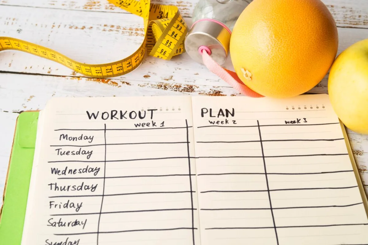 Open workout planner with a measuring tape, water bottle, and fruit on a wooden surface.