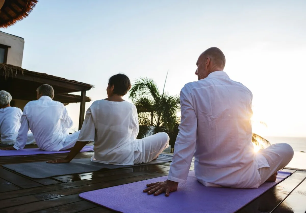 Group of adults practicing yoga outdoors at sunset, facing the sea on a wooden deck.