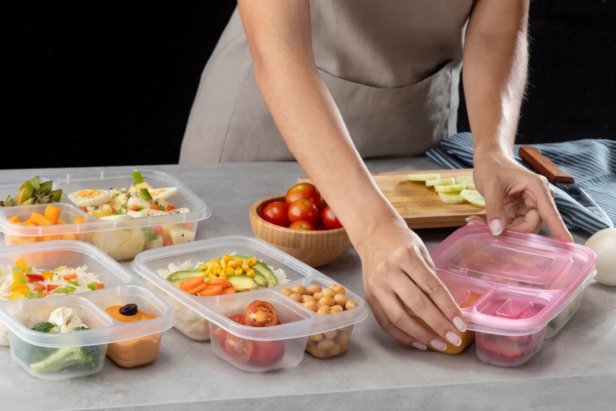 Person preparing various vegetables and placing them into multiple meal prep containers on a kitchen counter.