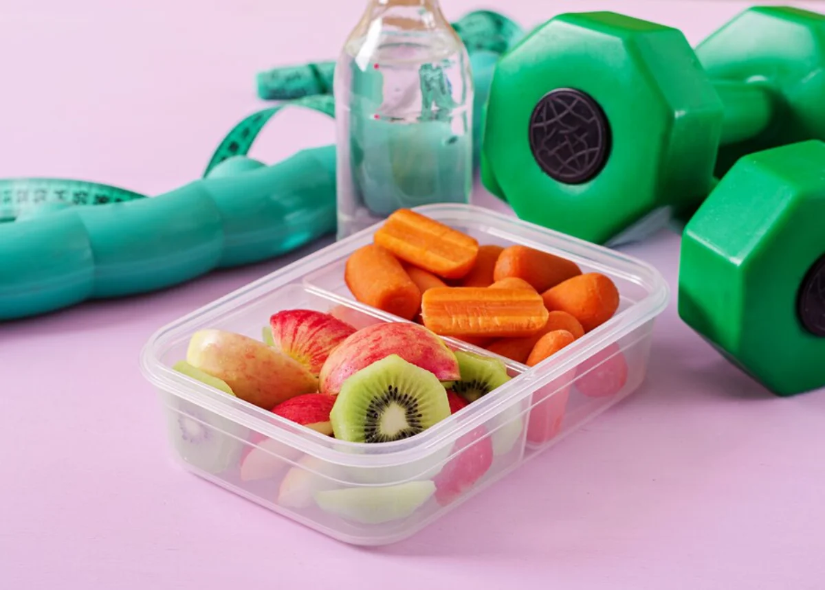 Healthy snacks in a clear container with dumbbells, water bottle, and measuring tape on a pink background.