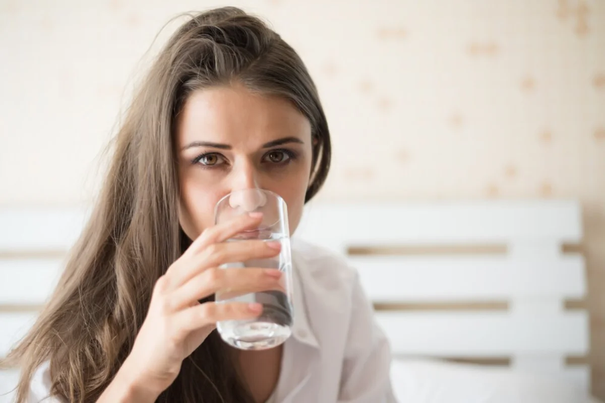 A woman in a white shirt drinking a glass of water indoors.