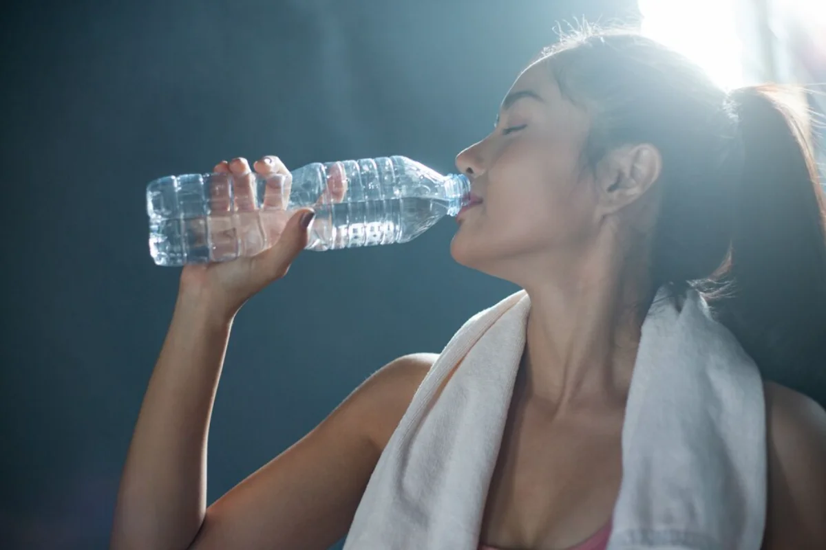 Woman drinking water from a bottle after workout, with a towel around her neck.