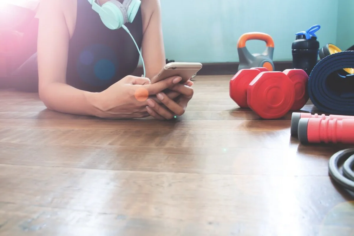 Woman using smartphone in gym with headphones and exercise equipment on wooden floor.
