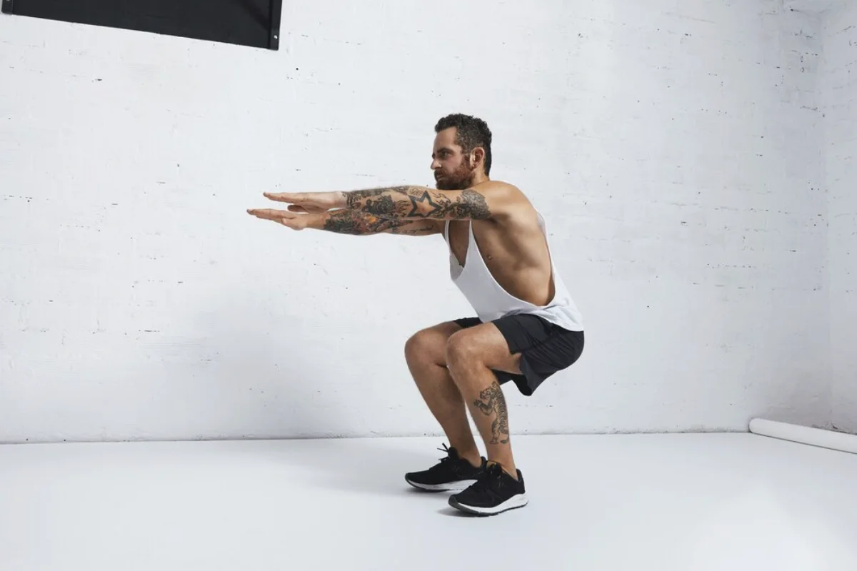 A man with tattoos performs a squat exercise in a minimalist studio setting.