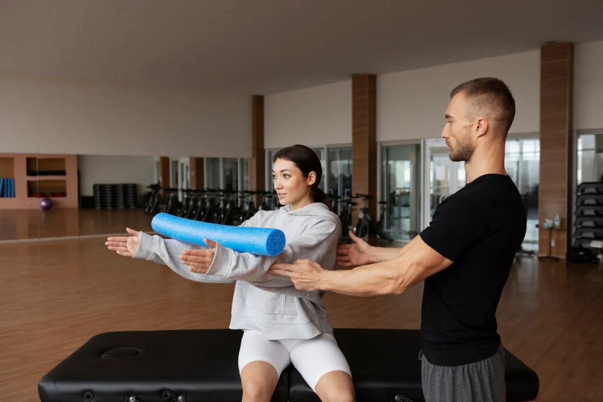 A man assists a woman with a stretching exercise using a foam roller in a well-equipped gym.