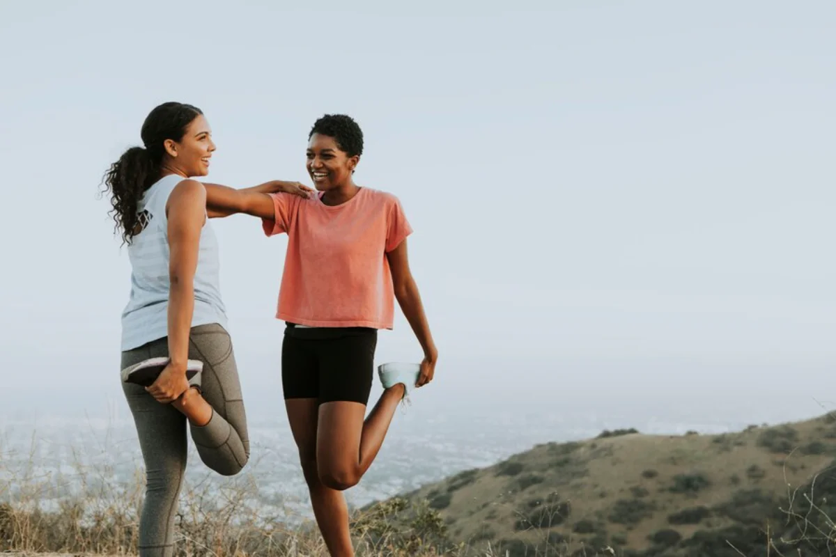 Two women stretching outdoors, smiling and enjoying a workout in a hilly landscape.