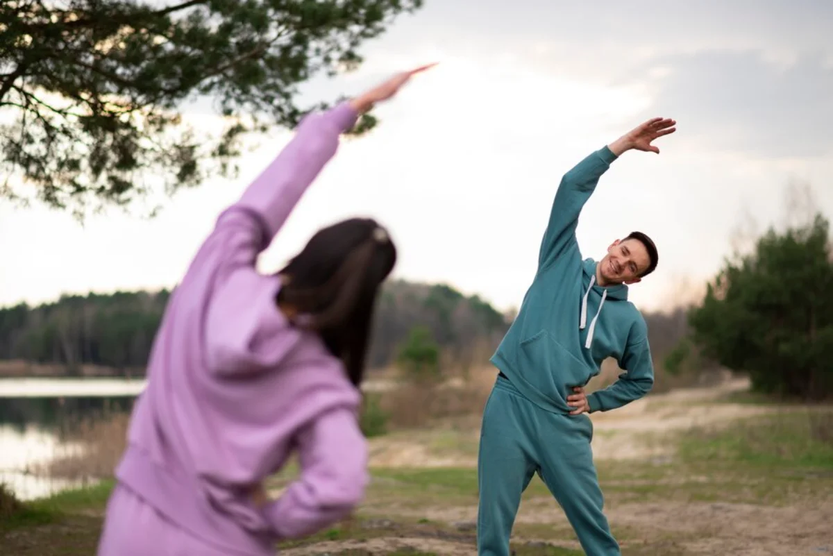 Two people in tracksuits stretching outdoors near a lake and pine trees.