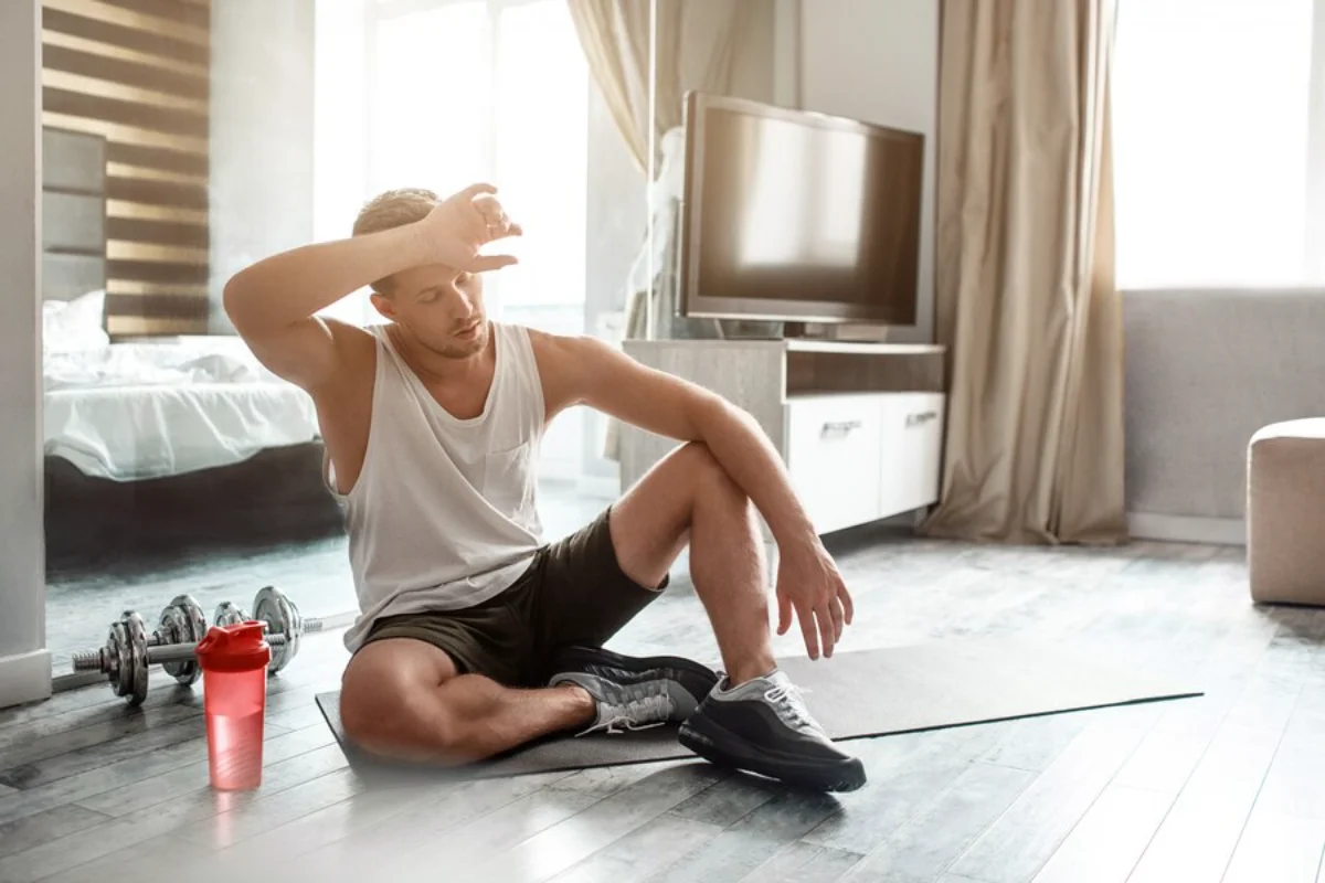 Man wiping sweat off his forehead after a workout, sitting next to weights on a living room floor.