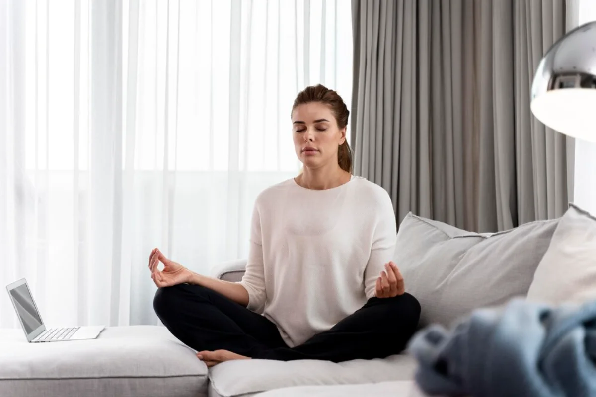 Woman meditating on bed in a bright room with a laptop nearby, promoting relaxation and mindfulness.