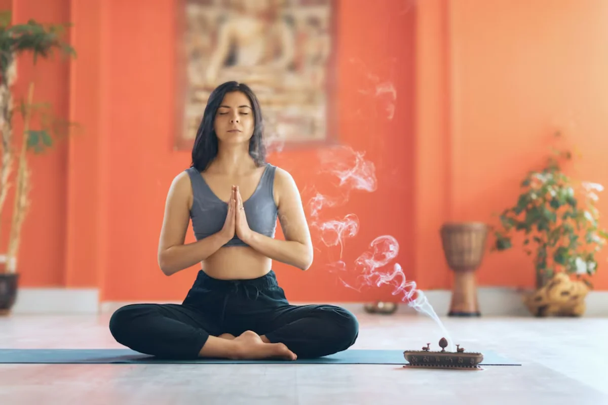 Woman meditating in lotus position with incense in a room with orange walls.