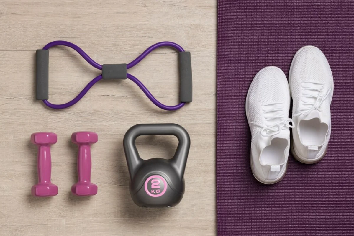 Top view of exercise equipment including white sneakers, purple resistance band, pink dumbbells, and a black kettlebell on a wooden floor.