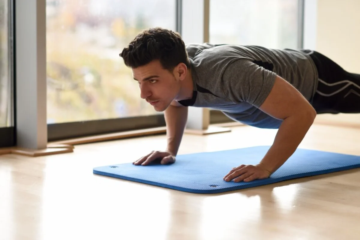 Man doing a plank exercise on a blue mat in a bright room near a window.