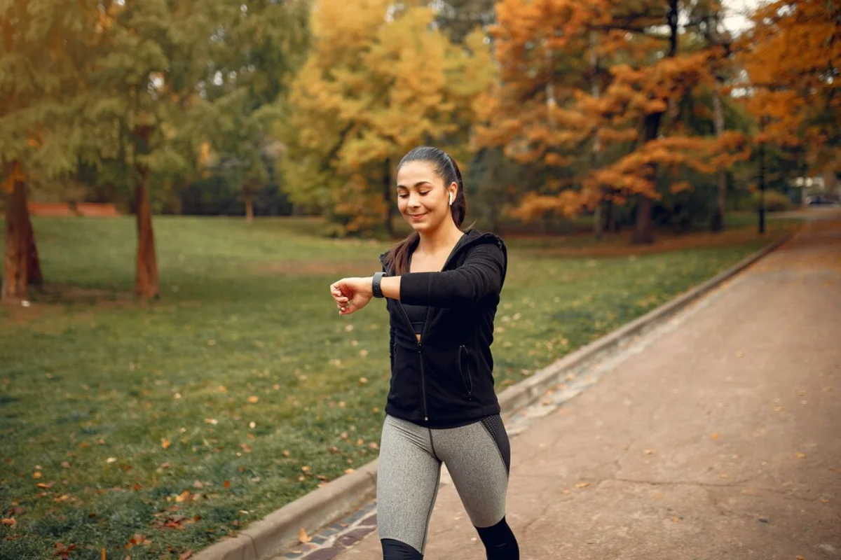 Woman checking her watch while walking in a park with autumn trees.