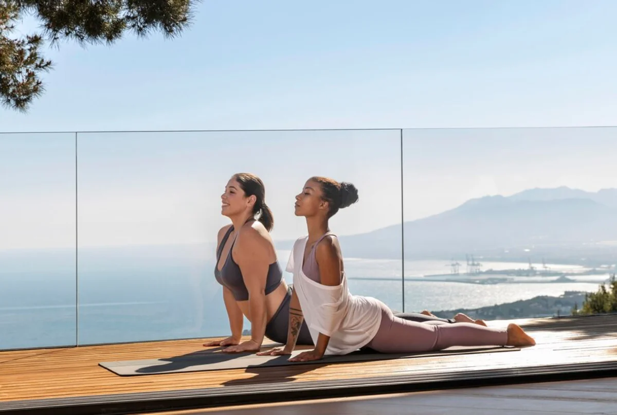 Two women practicing yoga on an outdoor deck overlooking the sea and mountains.