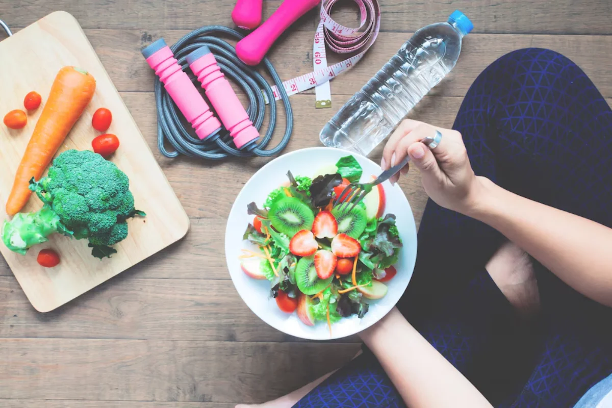 Top view of a person eating a healthy salad, with fitness equipment and fresh vegetables on a wooden table.