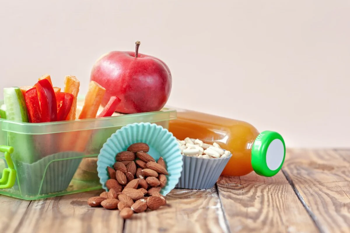 Healthy snacks on a table, including a red apple, carrot sticks, red pepper, almonds, and a juice bottle.