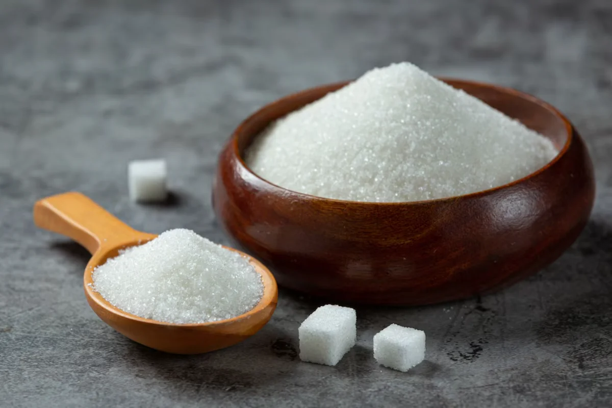 Granulated sugar in a wooden bowl and spoon, with sugar cubes on a grey surface.