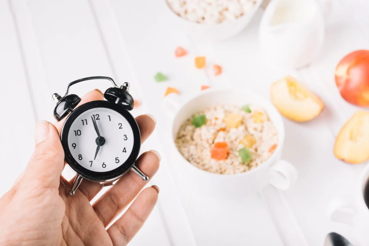 Hand holding a black alarm clock with a blurred breakfast setup in the background.
