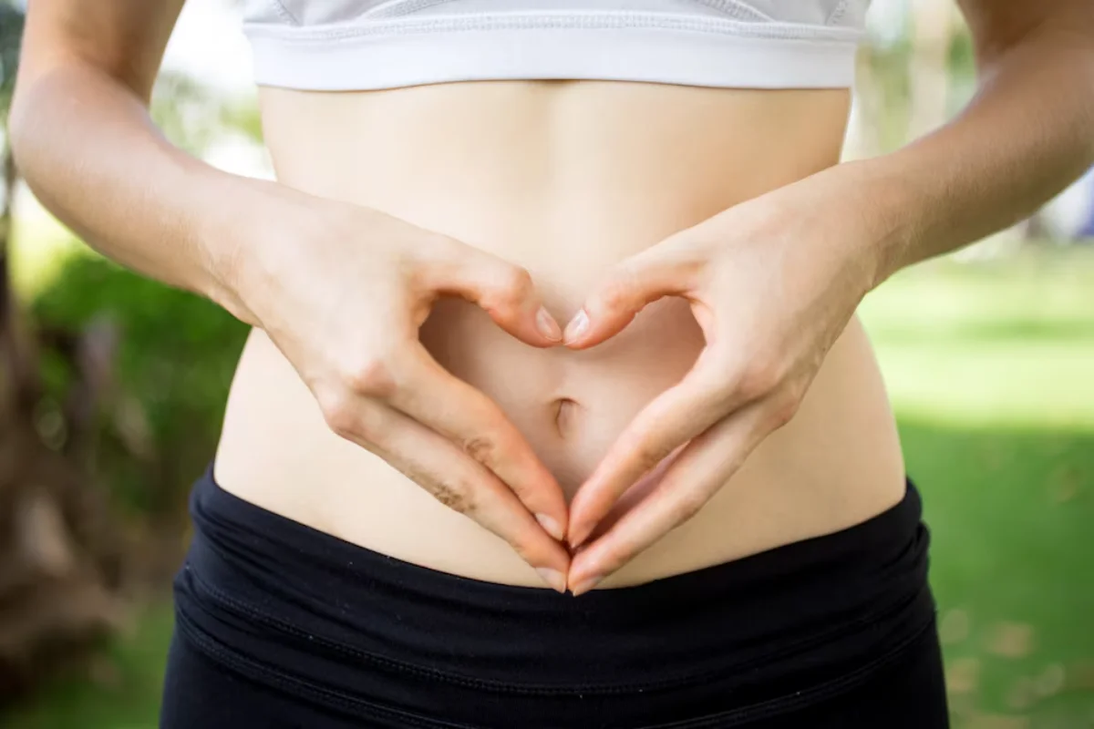 Close-up of a woman in a white crop top forming a heart shape with her hands around her stomach.